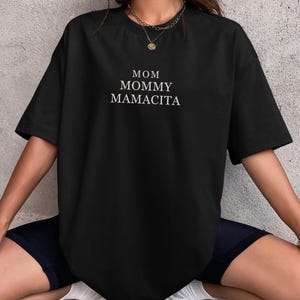 Pu&ograve; includere: T-shirt nero a maniche corte con la scritta bianca "MOM MOMMY MAMACITA". La maglietta &egrave; di taglio ampio. La modella indossa pantaloncini neri e scarpe da ginnastica bianche.