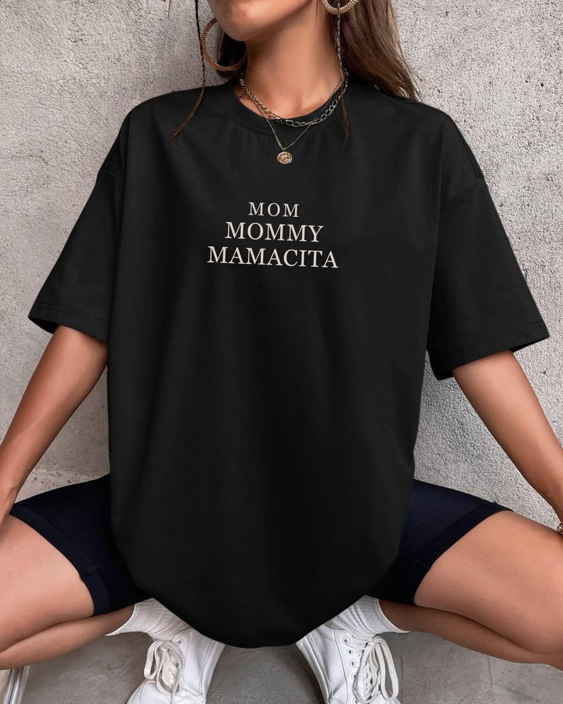 Pu&ograve; includere: T-shirt nero a maniche corte con la scritta bianca "MOM MOMMY MAMACITA". La maglietta &egrave; di taglio ampio. La modella indossa pantaloncini neri e scarpe da ginnastica bianche.