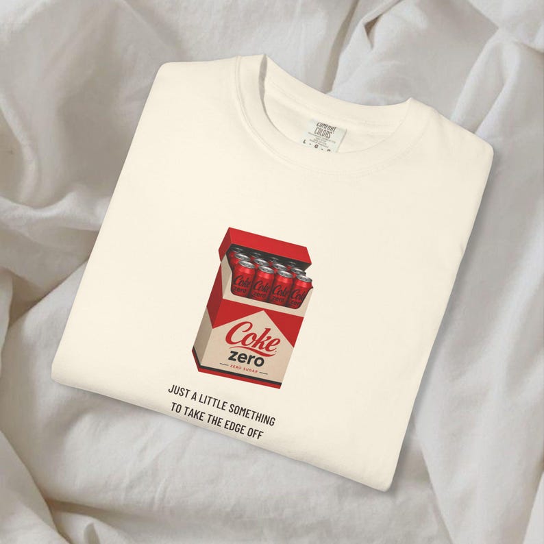 May include: A cream-colored t-shirt featuring a graphic of a red box containing red soda cans with the text 'Coke zero'. Below the graphic, the black text reads 'JUST A LITTLE SOMETHING TO TAKE THE EDGE OFF'. The shirt has a crew neckline.