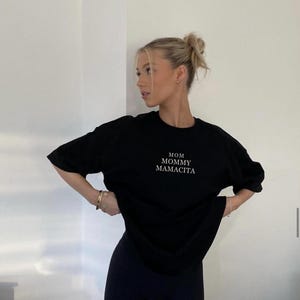Pu&ograve; includere: T-shirt nero con le parole "MOM MOMMY MAMACITA" in lettere bianche. Abbinato a pantaloncini sportivi neri. La modella indossa bracciali e orecchini dorati.