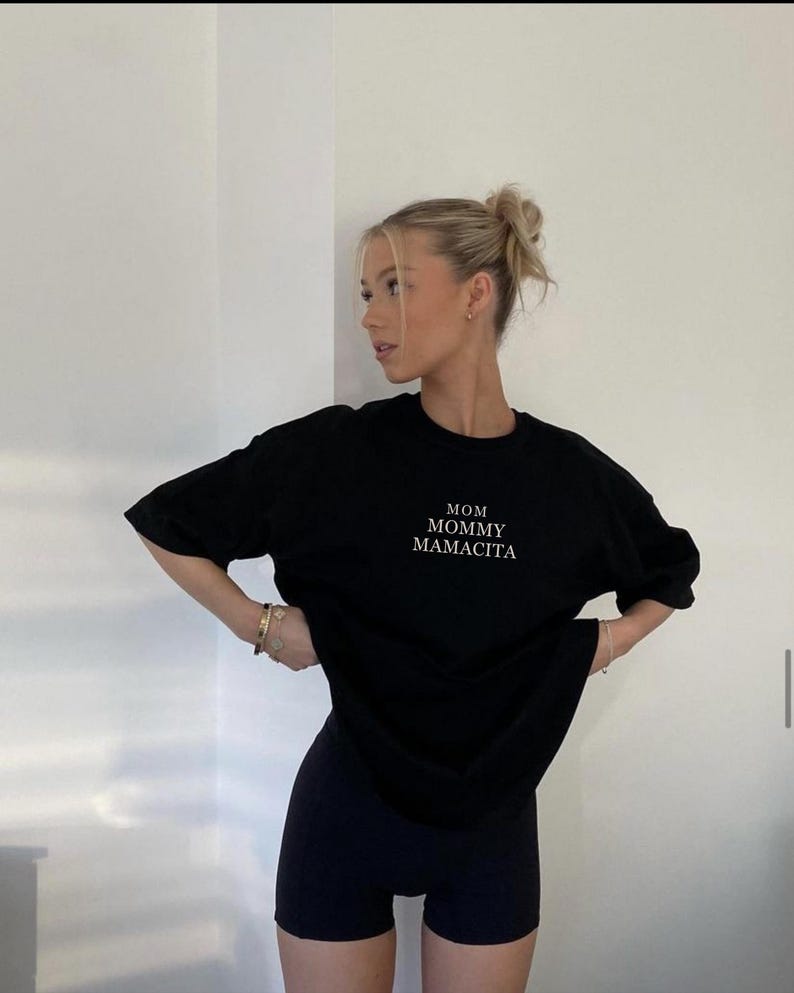 Pu&ograve; includere: T-shirt nero con le parole "MOM MOMMY MAMACITA" in lettere bianche. Abbinato a pantaloncini sportivi neri. La modella indossa bracciali e orecchini dorati.