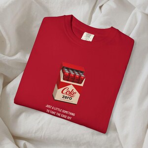 May include: A red t-shirt featuring a graphic of a cigarette-style pack filled with red Coke Zero soda cans. Below the graphic, the text reads: JUST A LITTLE SOMETHING TO TAKE THE EDGE OFF. The shirt has a crew neckline and a white interior brand label.