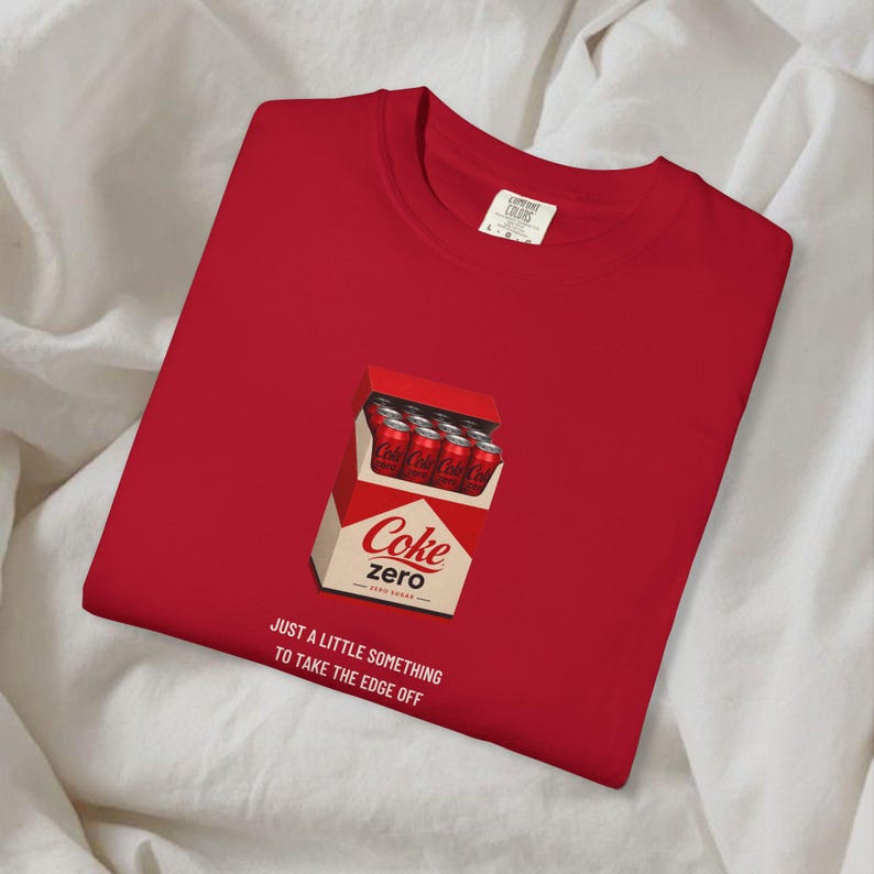 May include: A red t-shirt featuring a graphic of a cigarette-style pack filled with red Coke Zero soda cans. Below the graphic, the text reads: JUST A LITTLE SOMETHING TO TAKE THE EDGE OFF. The shirt has a crew neckline and a white interior brand label.