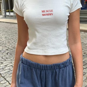 May include: A white, short-sleeved crop top with the red text "MUSCLE MOMMY". The top is worn with blue sweatpants. The outfit is casual and suitable for everyday wear.