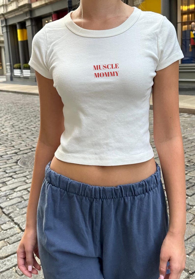 May include: A white, short-sleeved crop top with the red text "MUSCLE MOMMY". The top is worn with blue sweatpants. The outfit is casual and suitable for everyday wear.