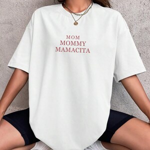 Pu&ograve; includere: T-shirt bianco a maniche corte con le parole "MOM MOMMY MAMACITA" stampate in rosso. La maglietta &egrave; in stile casual, adatta per l'uso quotidiano.
