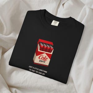 May include: A black t-shirt featuring a graphic of a red and white cigarette-style pack filled with red soda cans. The pack is labeled Coke zero. Below the graphic, the text reads: JUST A LITTLE SOMETHING TO TAKE THE EDGE OFF.