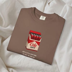 May include: A brown crew neck t-shirt featuring a graphic of a red and white Coke Zero carton filled with soda cans. Below the carton, the text reads: JUST A LITTLE SOMETHING TO TAKE THE EDGE OFF. The shirt has a white interior neck label.