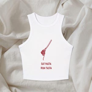 Puede incluir: Un top corto sin mangas de color blanco con un gráfico de líneas rojas de un tenedor enrollando espaguetis. Debajo del gráfico, el texto EAT PASTA RUN FASTA está impreso en letras mayúsculas rojas. La prenda tiene cuello alto y dobladillo corto.