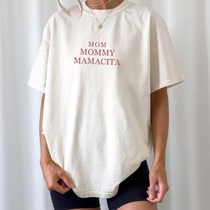Pu&ograve; includere: T-shirt color crema con le parole "MOM, MOMMY, MAMACITA" stampate in bordeaux. La maglietta ha una vestibilit&agrave; ampia e maniche corte. La modella indossa pantaloncini neri.