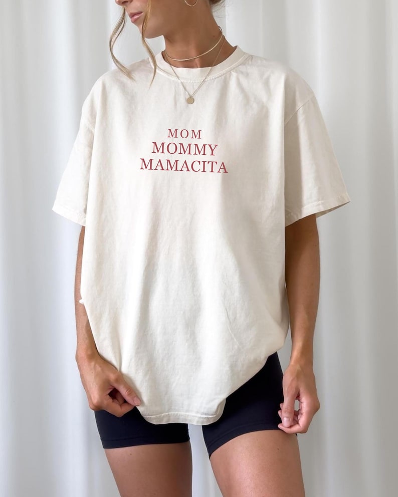 Pu&ograve; includere: T-shirt color crema con le parole "MOM, MOMMY, MAMACITA" stampate in bordeaux. La maglietta ha una vestibilit&agrave; ampia e maniche corte. La modella indossa pantaloncini neri.