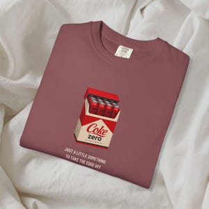 May include: A dusty rose crewneck t-shirt featuring a graphic of a red and white box containing red soda cans. The box is labeled Coke Zero. Below the graphic, the text reads: JUST A LITTLE SOMETHING TO TAKE THE EDGE OFF. The shirt has a size L tag.