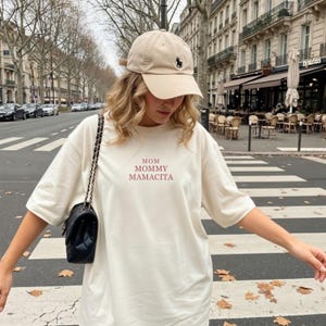 Pu&ograve; includere: T-shirt color crema con la scritta "MOM MOMMY MAMACITA" in bordeaux. La persona indossa un berretto da baseball beige con un logo nero e una borsa a tracolla nera. Lo sfondo mostra una via cittadina.