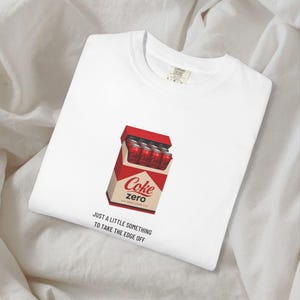 May include: A white t-shirt featuring a graphic of a red box containing multiple red soda cans with the text Coke zero. Below the graphic, the black text reads: JUST A LITTLE SOMETHING TO TAKE THE EDGE OFF. The shirt has a crew neckline and a tag.
