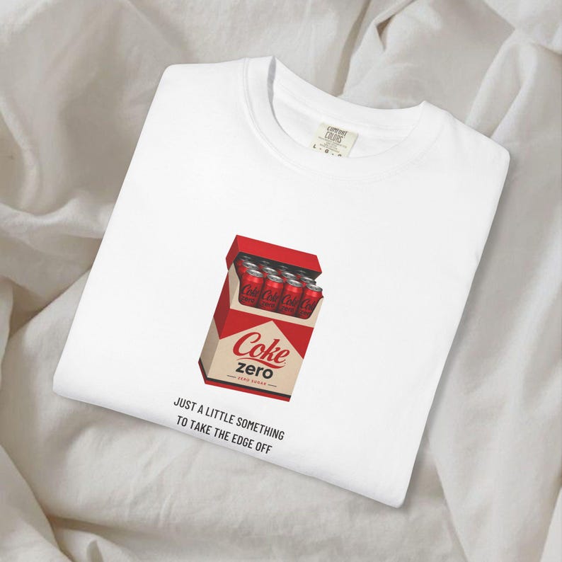 May include: A white t-shirt featuring a graphic of a red box containing multiple red soda cans with the text Coke zero. Below the graphic, the black text reads: JUST A LITTLE SOMETHING TO TAKE THE EDGE OFF. The shirt has a crew neckline and a tag.