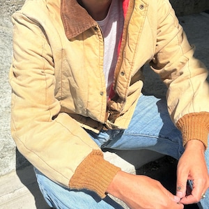 May include: A tan canvas jacket featuring a brown corduroy collar, ribbed brown knit cuffs, and a front zip fastening with snap buttons. The jacket is worn over a white t-shirt and paired with light blue denim jeans, showing a red inner lining.