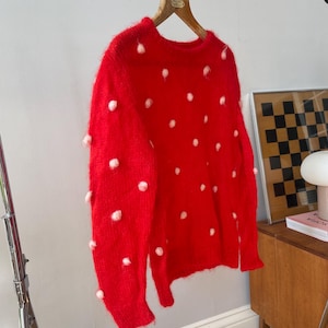 May include: A vibrant red long-sleeve sweater featuring a fuzzy, textured knit construction. The garment is adorned with scattered, small, pale pink pom-pom accents across the front and sleeves, finished with a classic crew neckline.