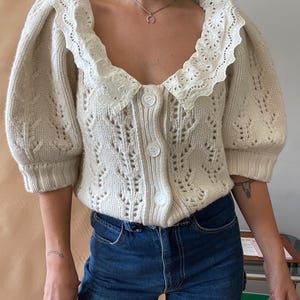 Op de afbeelding: Crèmekleurig vest met een geschulpte kanten kraag en pofmouwen. De trui met knopen heeft een delicaat open breiwerk. Het model draagt een blauwe jeans. Het vest is een stijlvolle toevoeging aan elke garderobe.