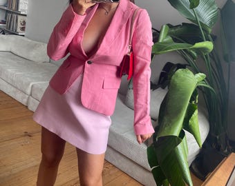 May include: A pink blazer with a single-button closure, paired with a matching pink mini skirt. A small red purse hangs from the shoulder. The outfit is completed with light blue boots. The background includes a couch and a large green plant.