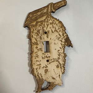 May include: A wooden light switch cover featuring a laser-engraved design of a possum carrying a log on its back. The possum is facing forward with its head tilted slightly to the side. The wood is a light brown color.