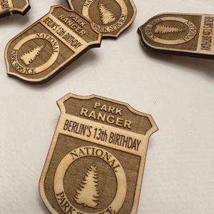 May include: A wooden park ranger badge with the text "Park Ranger" and "Berlin's 13th Birthday". The badge has a brown wood grain finish and features a circular design with a pine tree and the text "National Park Service".