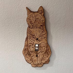 May include: Wooden light switch cover featuring a detailed engraving of a seated cat. The cat is depicted with intricate fur patterns and facial features. The light switch is a standard toggle style, and the cover is mounted on a white wall.