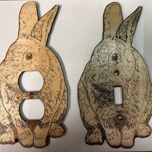 May include: Two wooden light switch and outlet covers featuring a laser-etched design of a rabbit. The rabbit is brown and white and has a realistic appearance.