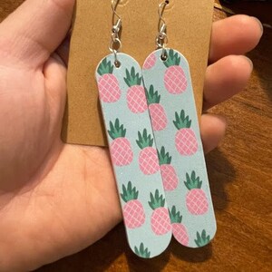 May include: A pair of long, light blue dangle earrings with a pink pineapple pattern. The earrings have silver hooks.