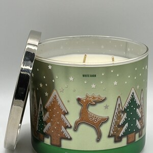 May include: A green and white candle jar with a silver lid. The jar has a festive design with gingerbread trees, a gingerbread reindeer, and white stars. The text "Fa La La Latte" and "White Barn" are printed on the jar. The text "Scented Luminary Candle Made With Natural Essential Oils" is printed on the bottom of the jar.
