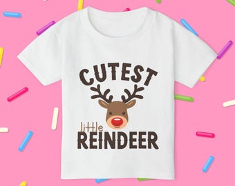 May include: White t-shirt with a reindeer graphic and the words "Cutest Little Reindeer" in brown. The reindeer has brown antlers, a red nose, and a smiling expression. The shirt is likely for a child.