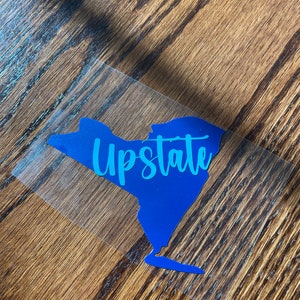 May include: A blue outline of the state of New York with the word "Upstate" written in teal inside the outline.
