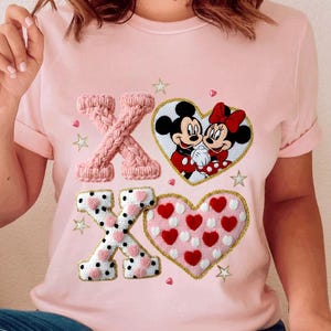 May include: A light pink t-shirt featuring a Valentine's Day design. The design includes the letters "XO" in a knitted style, along with two heart-shaped graphics. One heart features Mickey and Minnie Mouse, and the other has red hearts.
