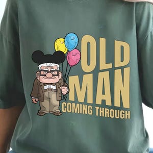Könnte beinhalten: Salbeigrünes T-Shirt mit einer Cartoon-Illustration eines alten Mannes mit Brille und schwarzem Hut, der bunte Luftballons hält. Der Text "OLD MAN COMING THROUGH" ist goldfarben gedruckt.