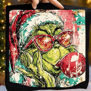 以下が含まれることがあります： 赤いサングラスとサンタ帽をかぶった緑色のグリンチのキャラクターが描かれた黒い正方形のクッション。 グリンチは赤いシャボン玉を吹いています。 背景は、赤、ティール、白の飛沫があるカラフルで使い古されたデザインです。