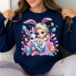 May include: Navy blue sweatshirt featuring a cartoon illustration of a princess holding a bunny with Easter eggs and pink splashes. The princess has blonde hair and blue eyes. The bunny is light purple with pink ears. The sweatshirt is a crew neck.