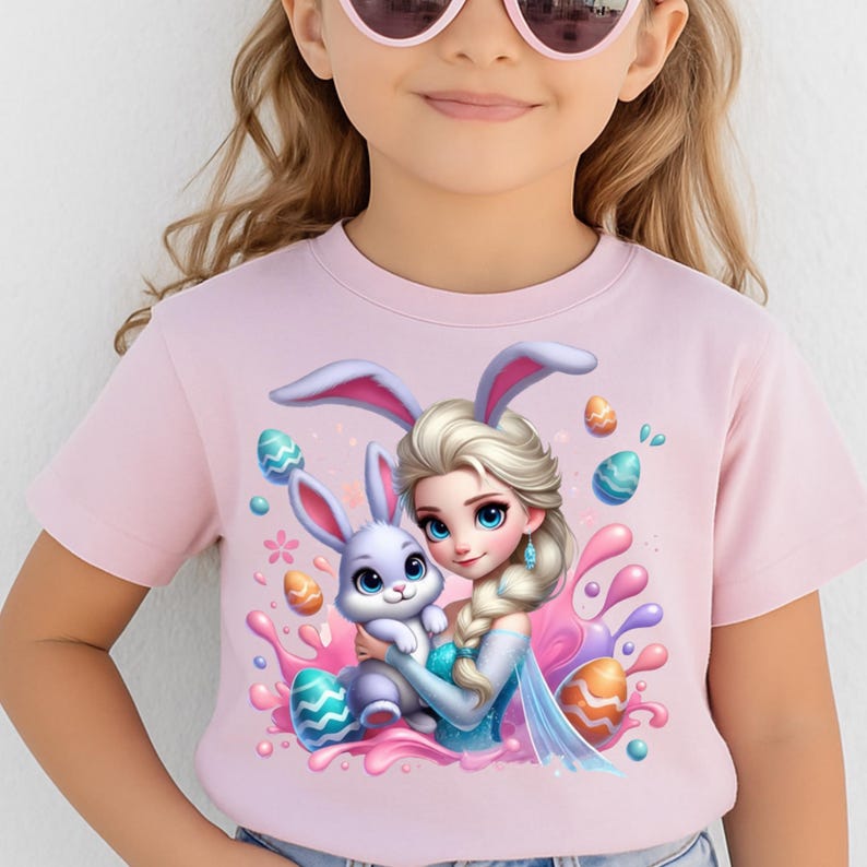 May include: A light pink t-shirt featuring a cartoon illustration of a princess with a bunny wearing bunny ears, surrounded by Easter eggs and colorful splashes. The princess is holding a small, light purple bunny.