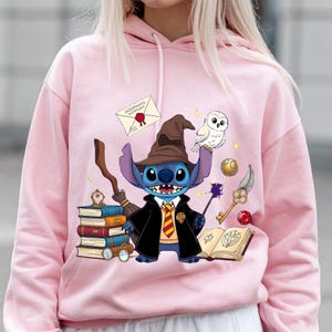 May include: Pink hoodie featuring a cartoon Stitch character dressed as a wizard, holding a wand and broom. The design includes a Hogwarts acceptance letter, books, an owl, and other magical elements.