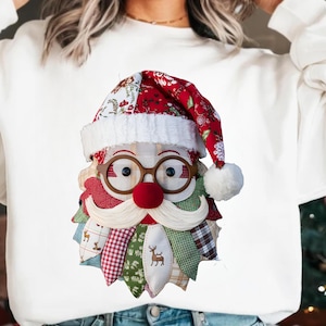 May include: White sweatshirt featuring a Santa Claus face design. The Santa face is made of fabric patches in red, green, and white, with a red nose, glasses, and a white beard. The Santa hat has a white pom-pom.