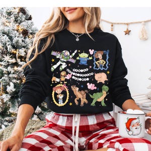 Puede incluir: Sudadera negra con personajes de dibujos animados coloridos y el texto "You've Got A Friend In Me". El dise&ntilde;o incluye im&aacute;genes de un guardabosques espacial, un extraterrestre, un vaquero y otros personajes. La sudadera se combina con pantalones de pijama a cuadros rojos y blancos.