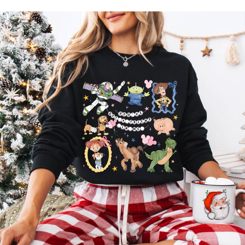 Puede incluir: Sudadera negra con personajes de dibujos animados coloridos y el texto "You've Got A Friend In Me". El dise&ntilde;o incluye im&aacute;genes de un guardabosques espacial, un extraterrestre, un vaquero y otros personajes. La sudadera se combina con pantalones de pijama a cuadros rojos y blancos.