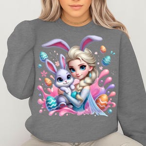 May include: Gray sweatshirt featuring a cartoon illustration of a princess with a bunny. The princess has blonde hair, blue eyes, and is wearing a blue dress. The bunny is light purple and is being held by the princess. Easter eggs and pink splashes surround the characters.