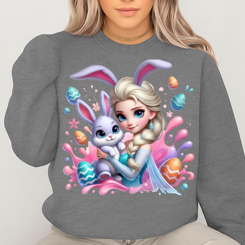 May include: Gray sweatshirt featuring a cartoon illustration of a princess with a bunny. The princess has blonde hair, blue eyes, and is wearing a blue dress. The bunny is light purple and is being held by the princess. Easter eggs and pink splashes surround the characters.