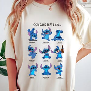 May include: Cream-colored t-shirt featuring a blue cartoon character in various poses, with the text "GOD SAYS THAT I AM..." above the images. Each image has a corresponding word like "EMPOWERED" and a Bible verse reference.