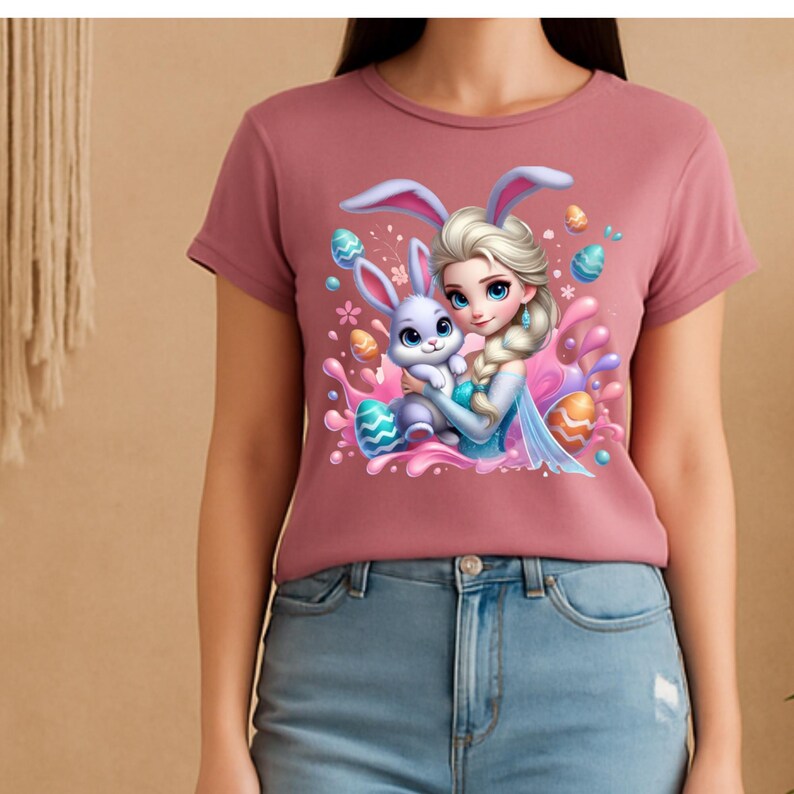May include: A mauve-colored t-shirt featuring a cartoon illustration of a princess with blonde hair holding a purple bunny with bunny ears. Easter eggs and pink splashes surround the characters. The shirt is paired with blue jeans.