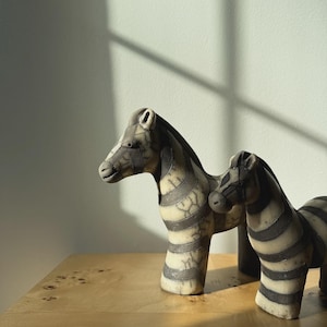 May include: Two decorative zebra figurines with black and white striped patterns. The zebras are standing on a wooden surface. The figurines have a textured appearance and are positioned in front of a light-colored wall.