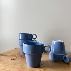 May include: Several blue ceramic mugs with circular handles are displayed on a light wooden surface. The mugs are stacked and arranged in a group. The mugs have a simple, modern design and a glossy finish. The background is a plain white wall.