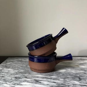 May include: Two stacked ceramic soup bowls. The bowls are brown with a dark blue rim and handle. The bowls are on a marble surface.