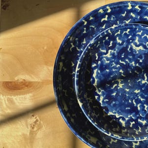 Puede incluir: Dos platos de cerámica azules con un patrón moteado blanquecino. Están apilados sobre una superficie de madera clara, con una sombra proyectada. Los platos son probablemente para servir alimentos.