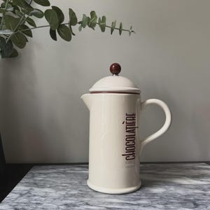 May include: Cream ceramic French press with a brown lid and handle. The text "la Chocolatière" is printed vertically on the side in brown. The press is on a marble surface with greenery in the background.