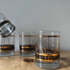 May include: Set of four clear glass tumblers with a black and gold geometric design. The glasses have a cylindrical shape with a thick gold band at the top and bottom, and vertical gold lines. One glass is tilted, showing the interior.